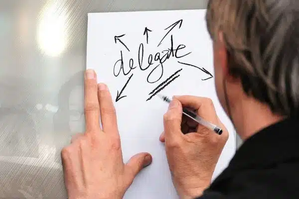 Hand writing the word delegate on a white paper with arrows pointing outward, signifying delegation