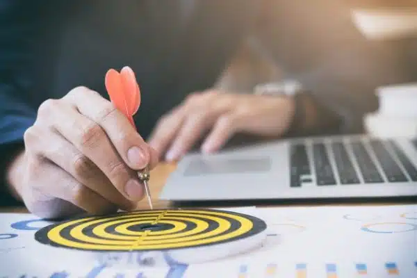 Hand holding a dart aimed at the bullseye of a yellow and black target, with a laptop in the background