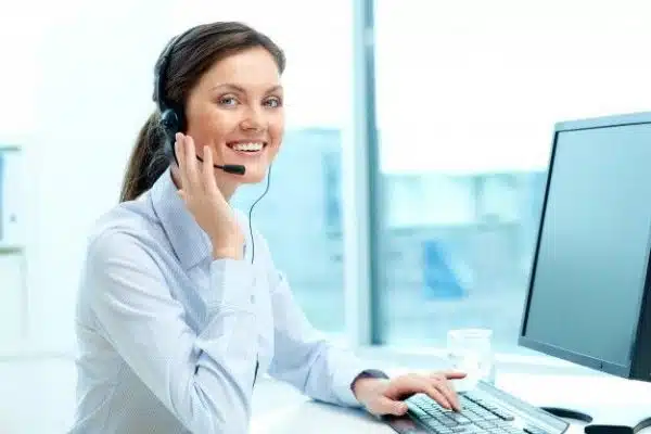 Woman in a white shirt wearing a headset, smiling, and sitting at a desk with a computer