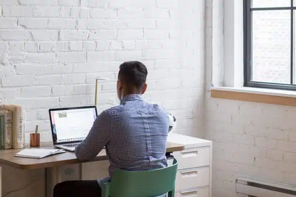 A man is working on his laptop at the table in a room, with a typewriter beside him.