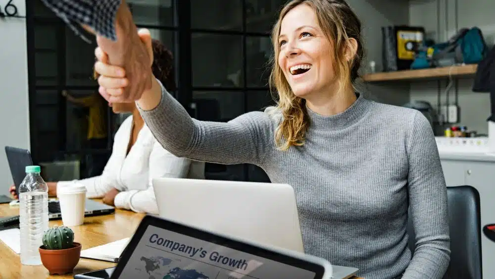 A woman smiles as she shakes hands with someone in an office setting.