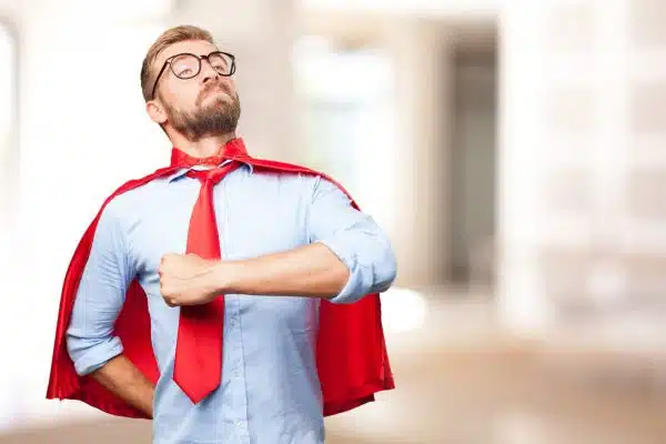 A confident man wearing glasses and a red superhero cape over a blue shirt and red tie, posing with a proud expression in an office setting