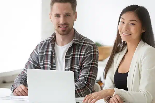 Two people, a man and a woman, sitting at a desk with a laptop. They are both smiling and looking at the camera