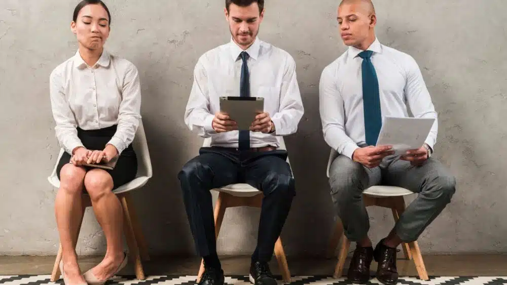 Three people sitting in a row on chairs against a plain background. The woman on the left looks unsure, the man in the middle is focused on a tablet, and the man on the right is glancing at a document