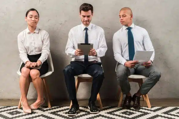 Three people sitting in a row on chairs against a plain background. The woman on the left looks unsure, the man in the middle is focused on a tablet, and the man on the right is glancing at a document