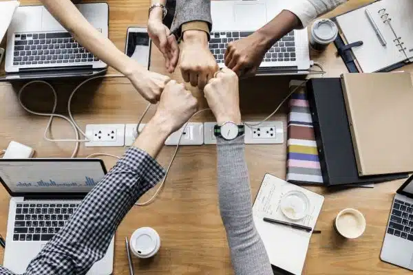 A group of people sitting around a wooden table, each with a laptop in front of them, putting their fists together in the center of the table in a gesture of teamwork